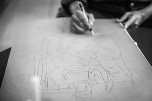 A black and white image of a man focused on drawing on a sheet of paper, showcasing his artistic concentration.