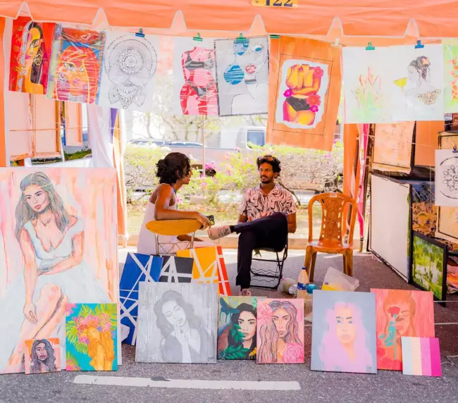 A woman seated at a table, surrounded by her vibrant paintings, showcasing her artistic talent and creativity.