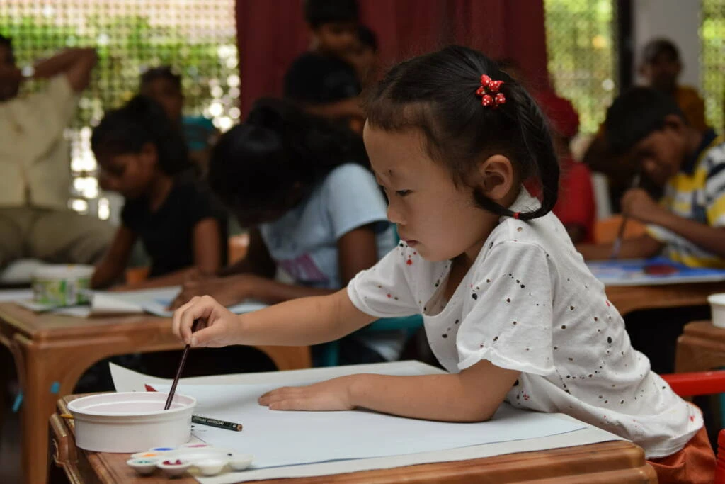 A little girl focused on painting at a table in a bright classroom, surrounded by art supplies and colorful decorations.