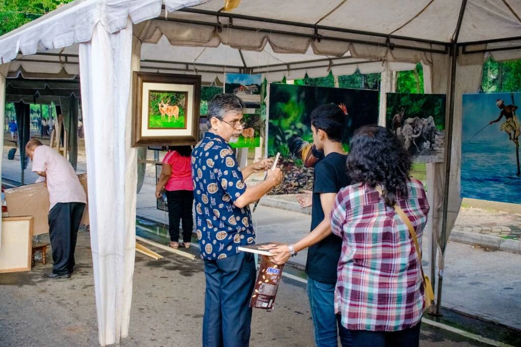 A diverse group of people admiring various paintings at an outdoor art fair, surrounded by vibrant displays and natural scenery.