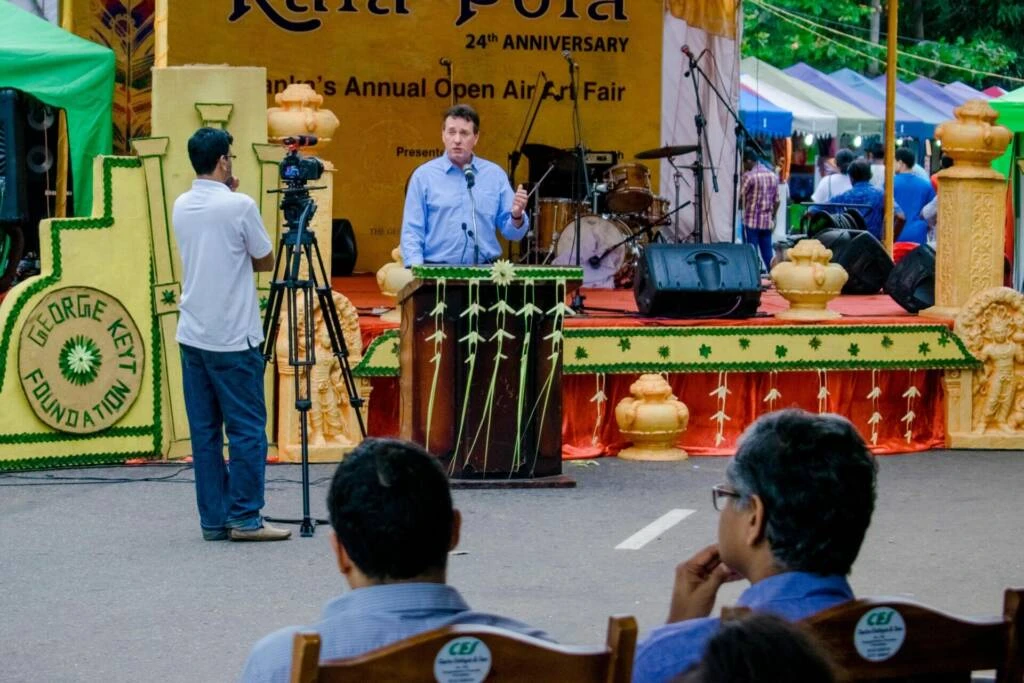 A man presents a speech on stage, with an attentive crowd watching and listening intently from the audience.