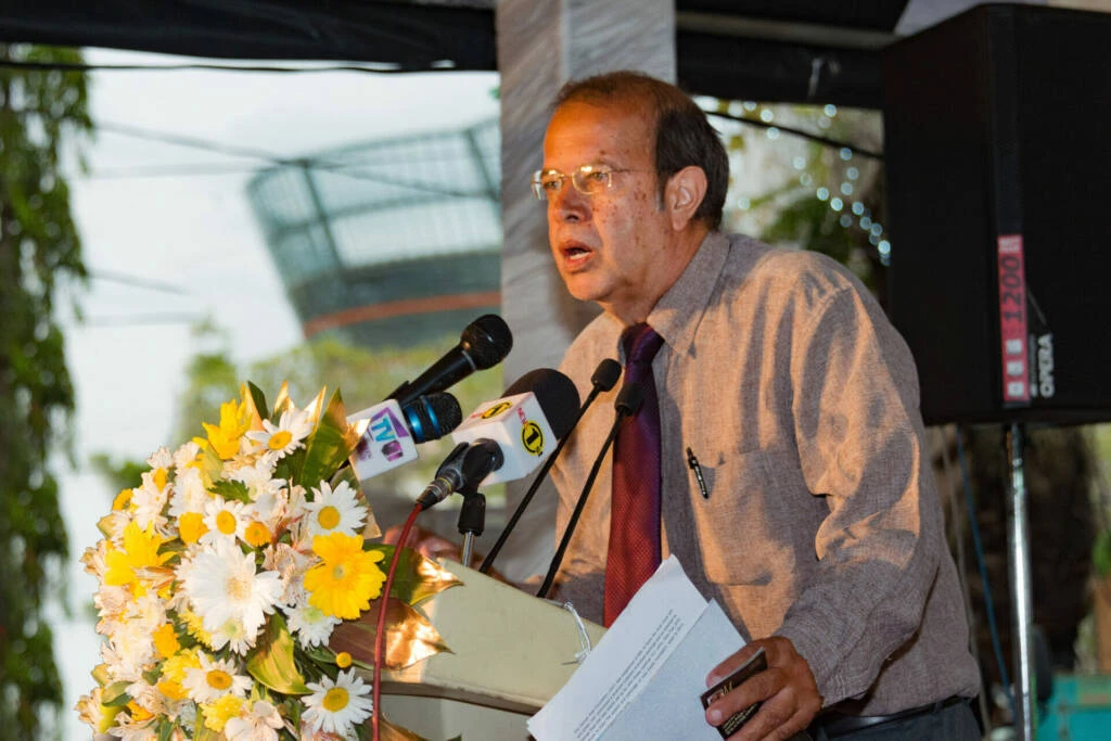 A man in a tie and glasses stands at a podium, addressing an audience with a speech.