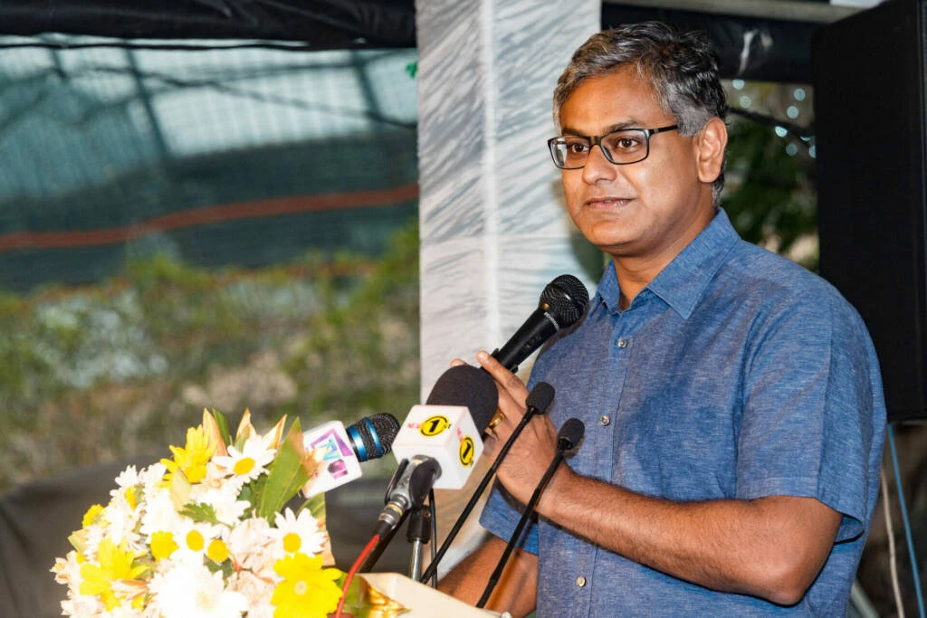 A man wearing glasses stands at a podium, delivering a speech to an audience with a focused expression.