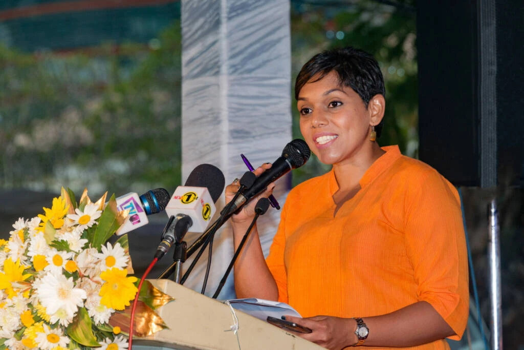 A woman wearing an orange shirt is speaking into a microphone, engaging her audience with confidence.