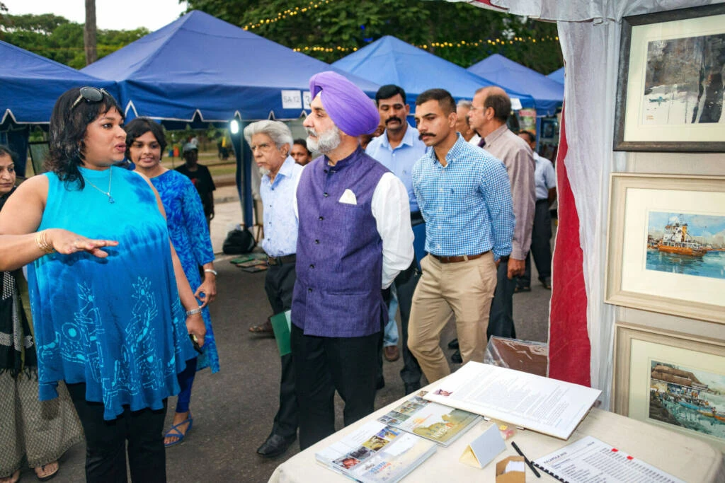 A man and woman stand beside a table displaying various paintings, engaged in conversation about the artwork.