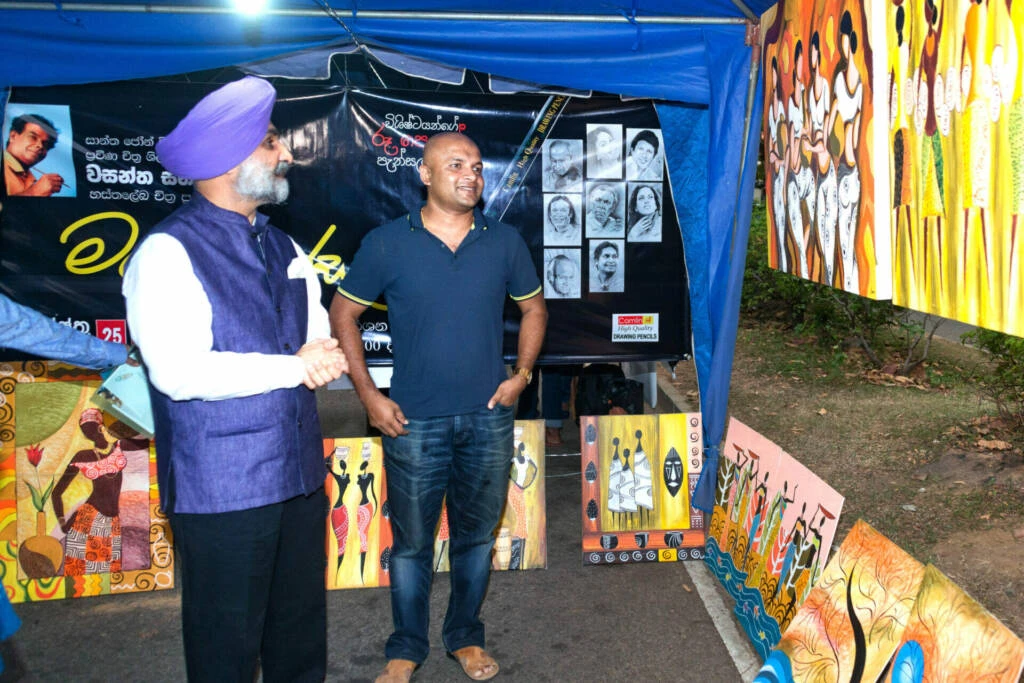 Two men stand beside various paintings displayed in an art gallery, engaging in conversation about the artwork.