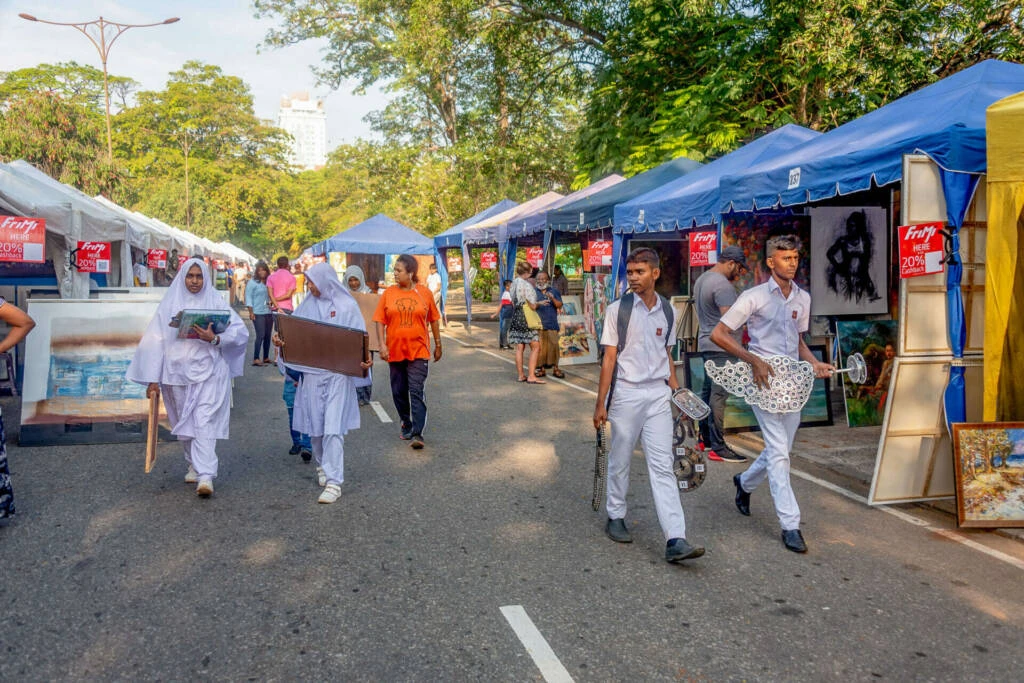 Two men examine various paintings displayed at an outdoor market, engaged in conversation about the artwork.