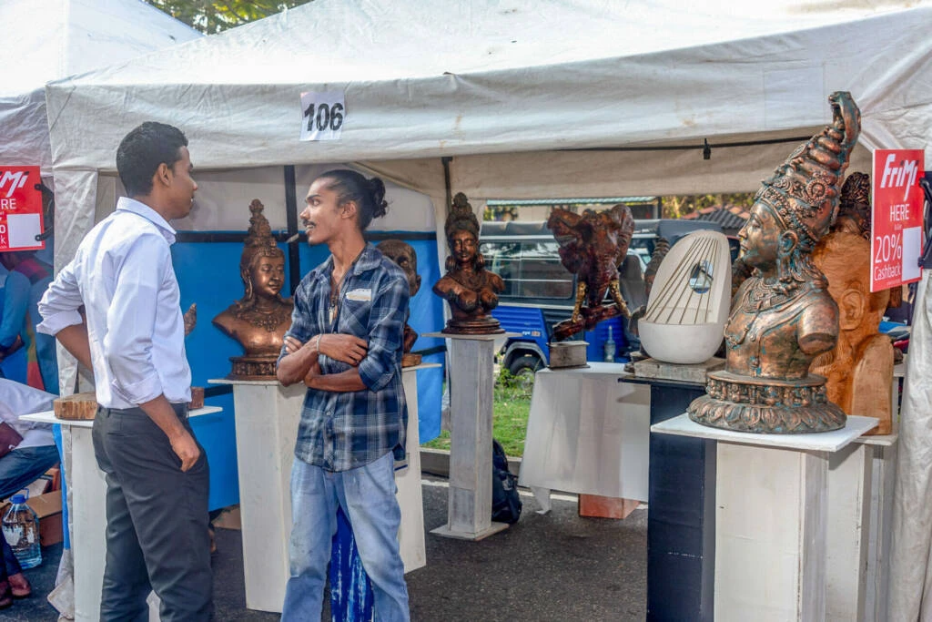 A man engages in conversation with a woman at a bustling outdoor market, surrounded by various stalls and shoppers.