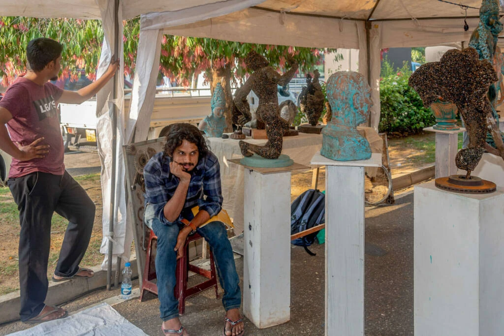 A man and a woman converse at an outdoor market, with colorful stalls and shoppers creating a lively atmosphere around them.