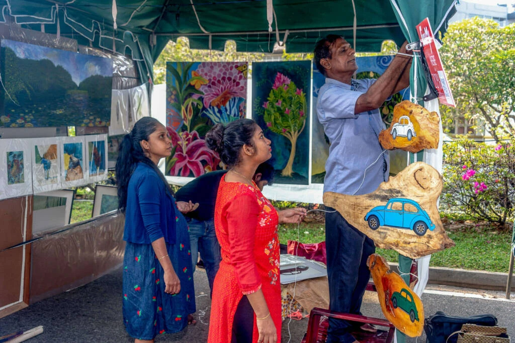 A man signs documents while two women stand beside him in front of tents, indicating a collaborative outdoor setting.
