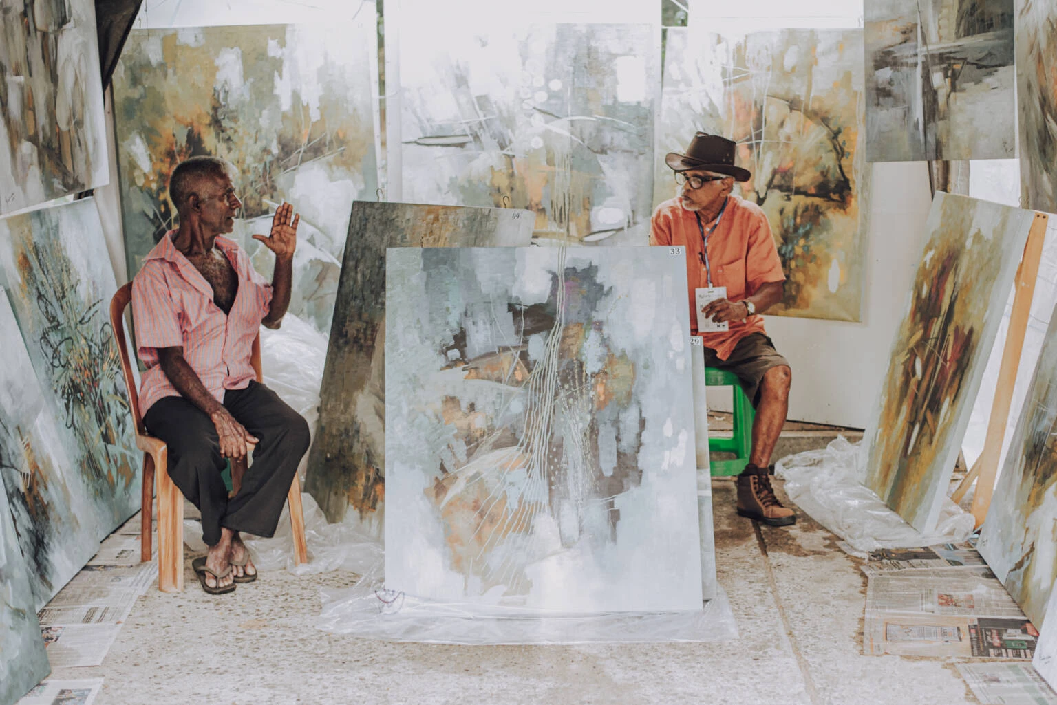 Two men seated in front of various paintings, engaged in conversation and appreciating the artwork displayed.