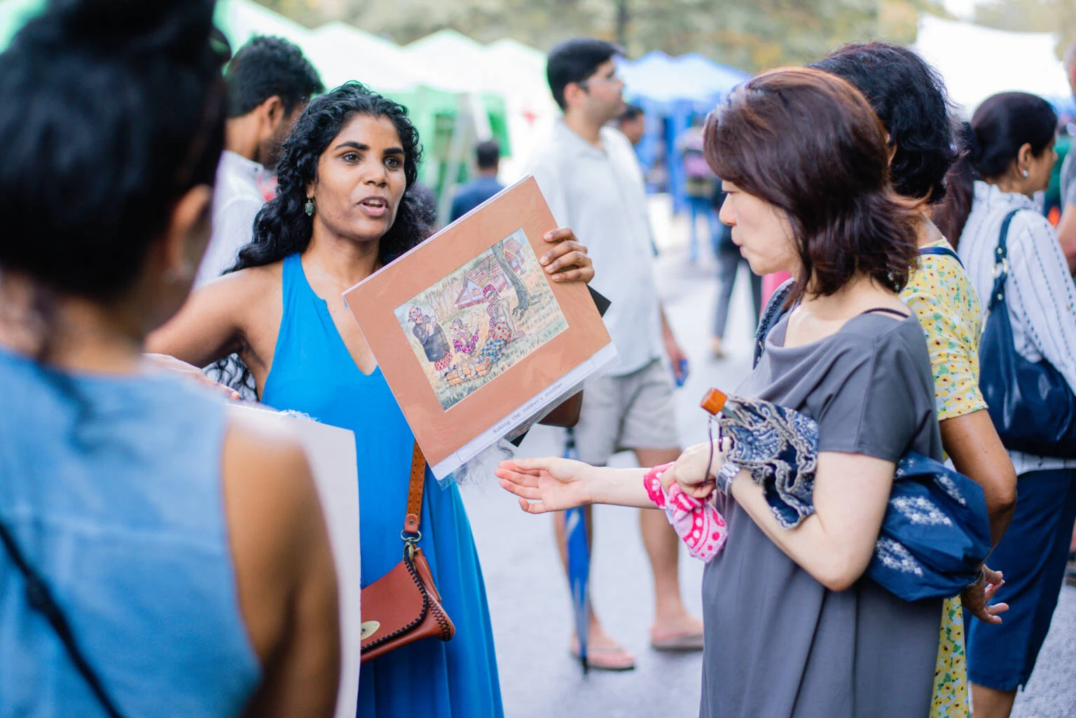 A woman engages in conversation with another woman at a bustling outdoor market, surrounded by various stalls and shoppers.