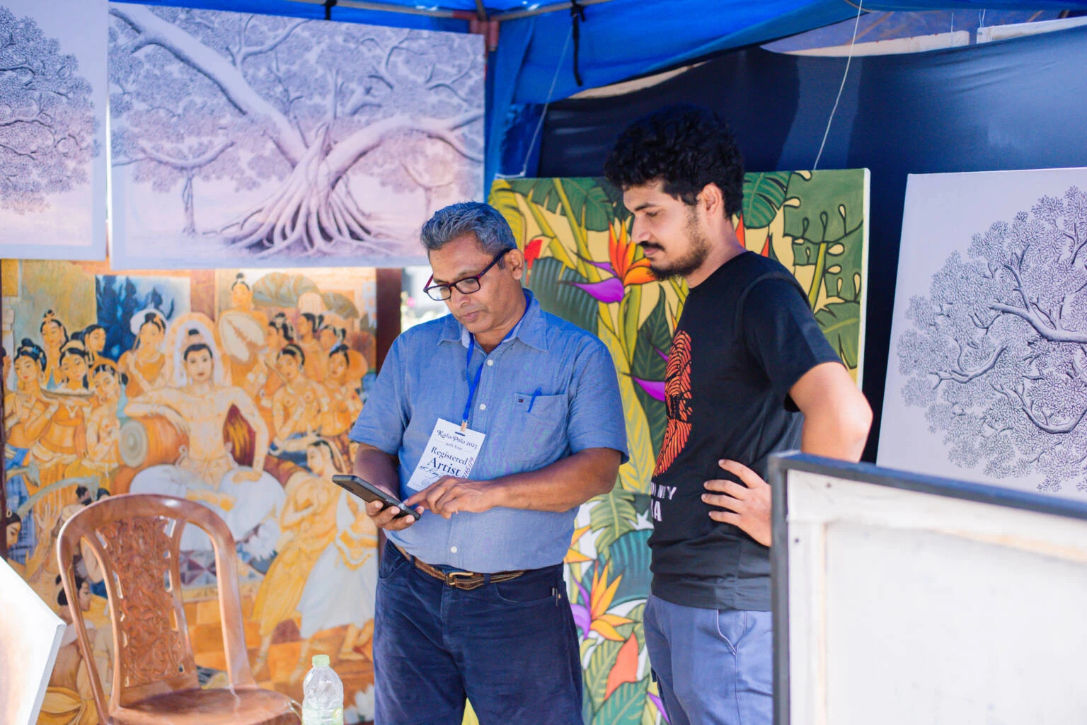 Two men stand side by side, one showing something on his phone to the other, both engaged in conversation.
