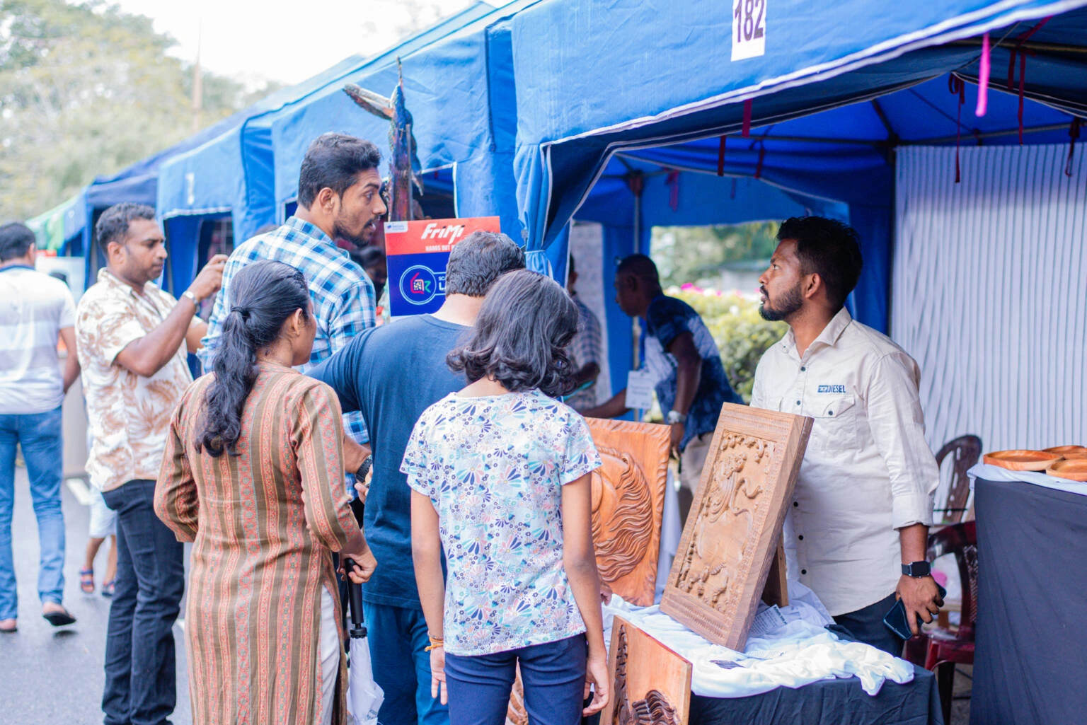 A group of people gathers around a booth where a man is selling wooden products, engaging in conversation and browsing items.