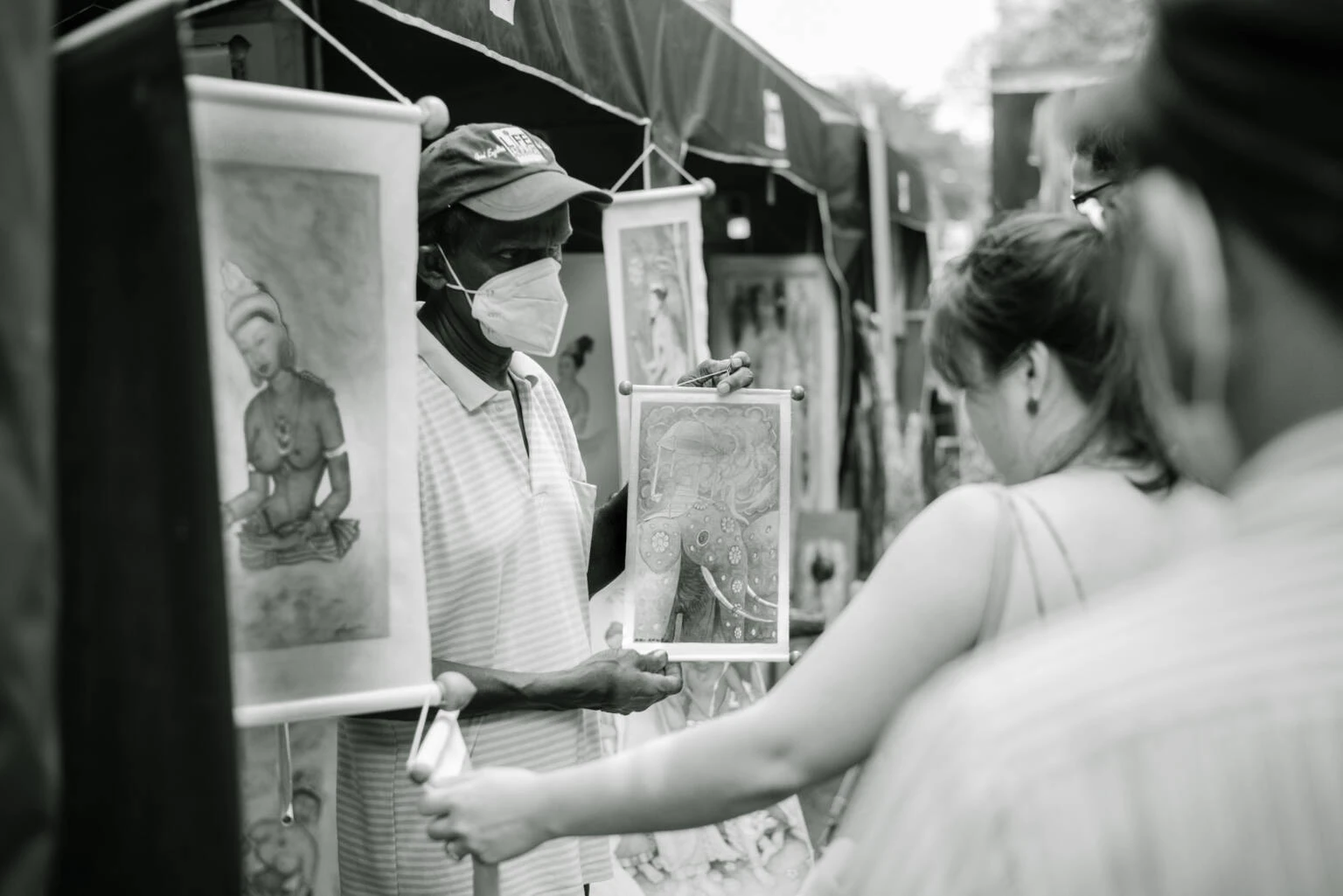A masked man stands beside a display of colorful paintings, engaging potential buyers in an outdoor setting.