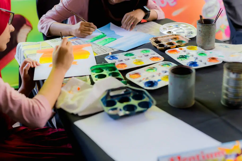 Two women collaboratively painting on paper at a table, showcasing creativity and teamwork in their artistic endeavor.
